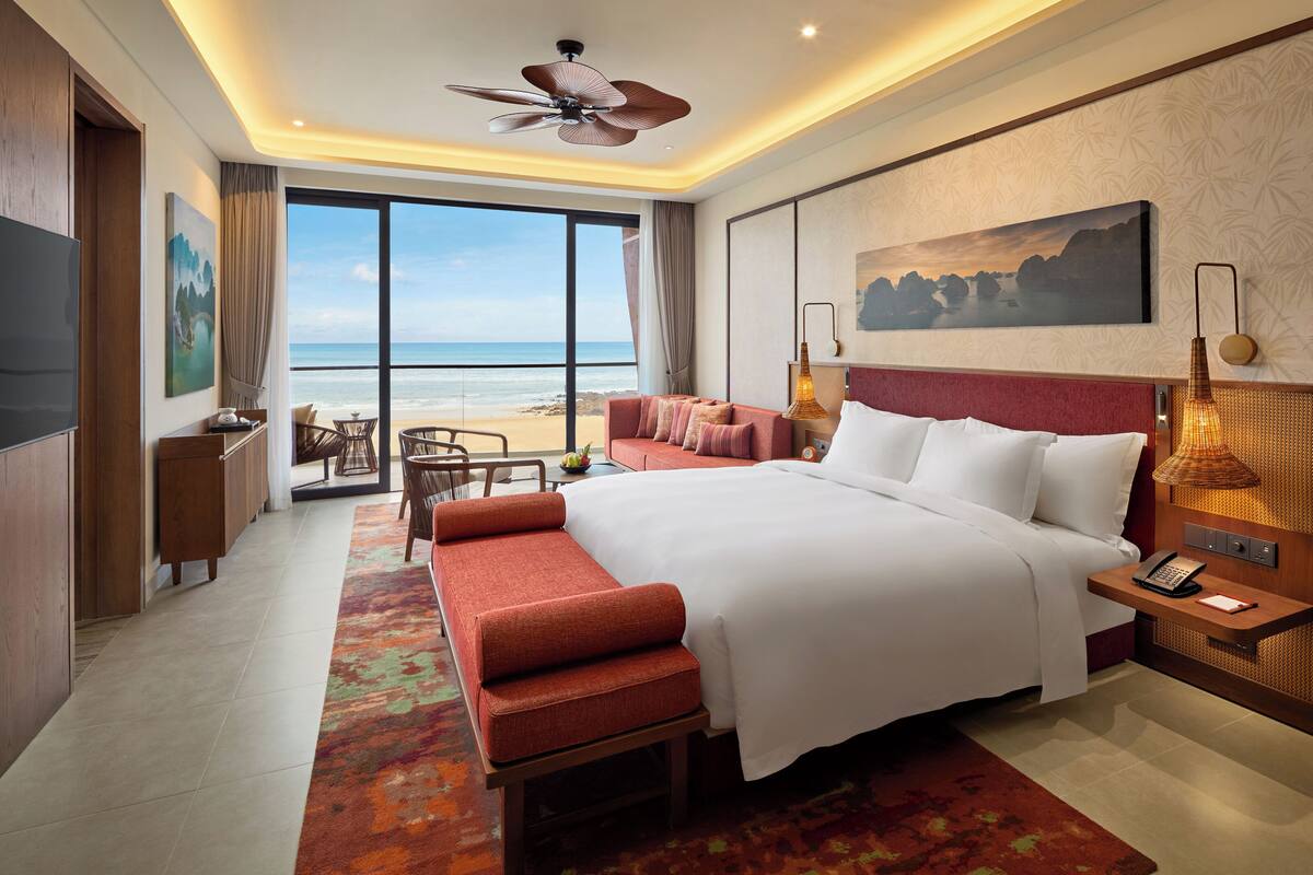 A hotel room with a large bed, a seating area with a view of the beach, a desk, and a TV.