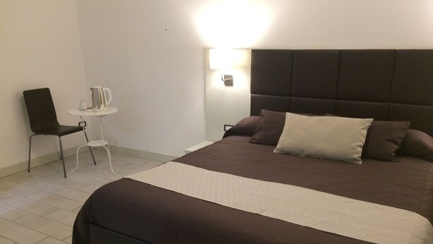 A neatly made bed with a dark headboard and multiple pillows, a small white table with a chair, and a wall-mounted light fixture. - Apeiron (Corigliano-Rossano)