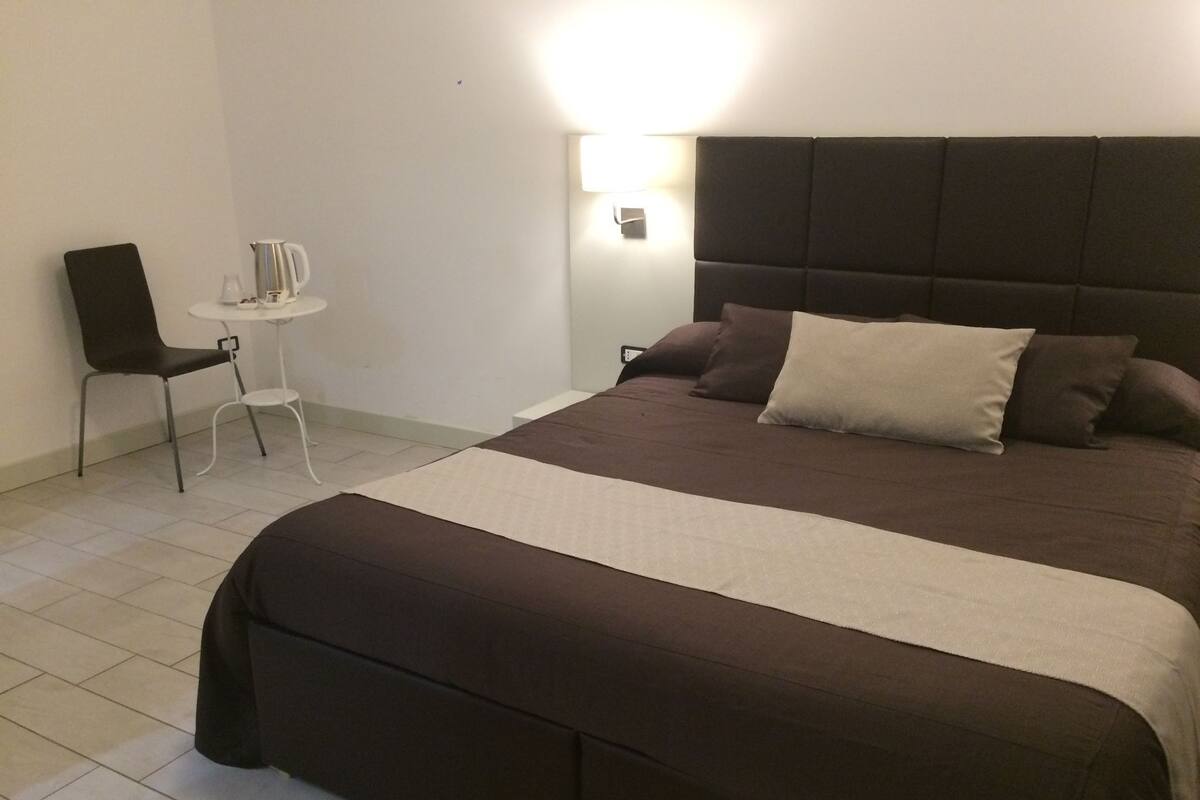 A neatly made bed with a dark headboard and multiple pillows, a small white table with a chair, and a wall-mounted light fixture.
