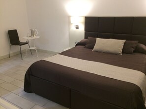 A neatly made bed with a dark headboard and multiple pillows, a small white table with a chair, and a wall-mounted light fixture. - Apeiron (Corigliano-Rossano)