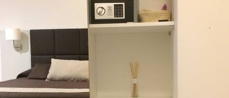 A bedroom with a bed, a bedside table with a microwave, a small refrigerator, and a wall-mounted lamp.