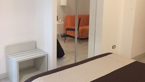 A hotel room with a bed, a small bedside table, a chair, a TV mounted on the wall, and a mirror. - Apeiron (Corigliano-Rossano)