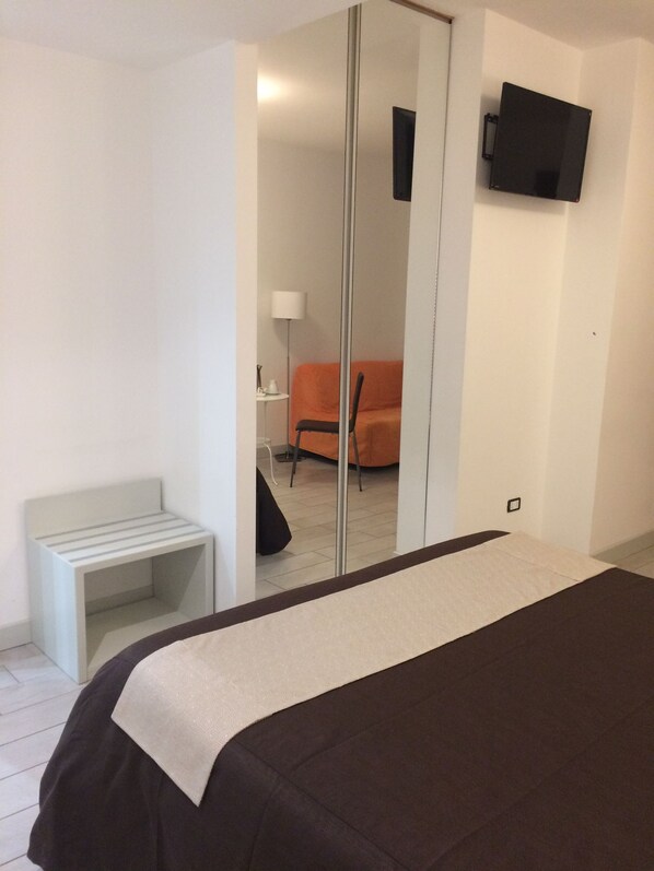 A hotel room with a bed, a small bedside table, a chair, a TV mounted on the wall, and a mirror. - Apeiron (Corigliano-Rossano)