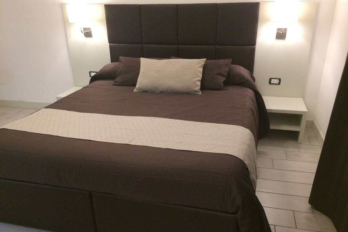 A neatly made bed with a dark headboard, two bedside tables, and a light-colored rug.