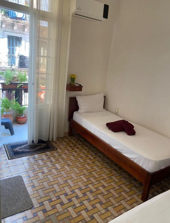 Una habitación pequeña con una cama individual, estructura de madera, colchón blanco y una mesita de noche con un florero.