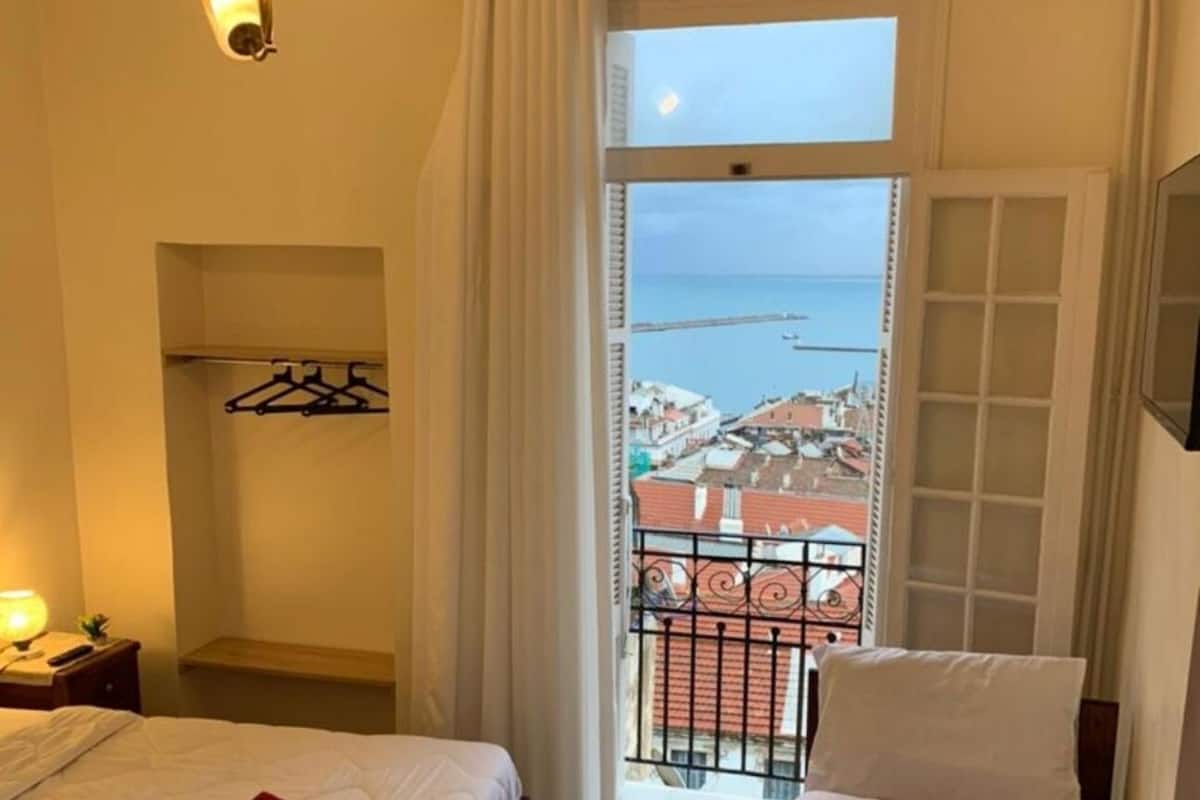 A hotel room with a bed, a nightstand, a hanging coat rack, and a balcony with a view of the sea and buildings.