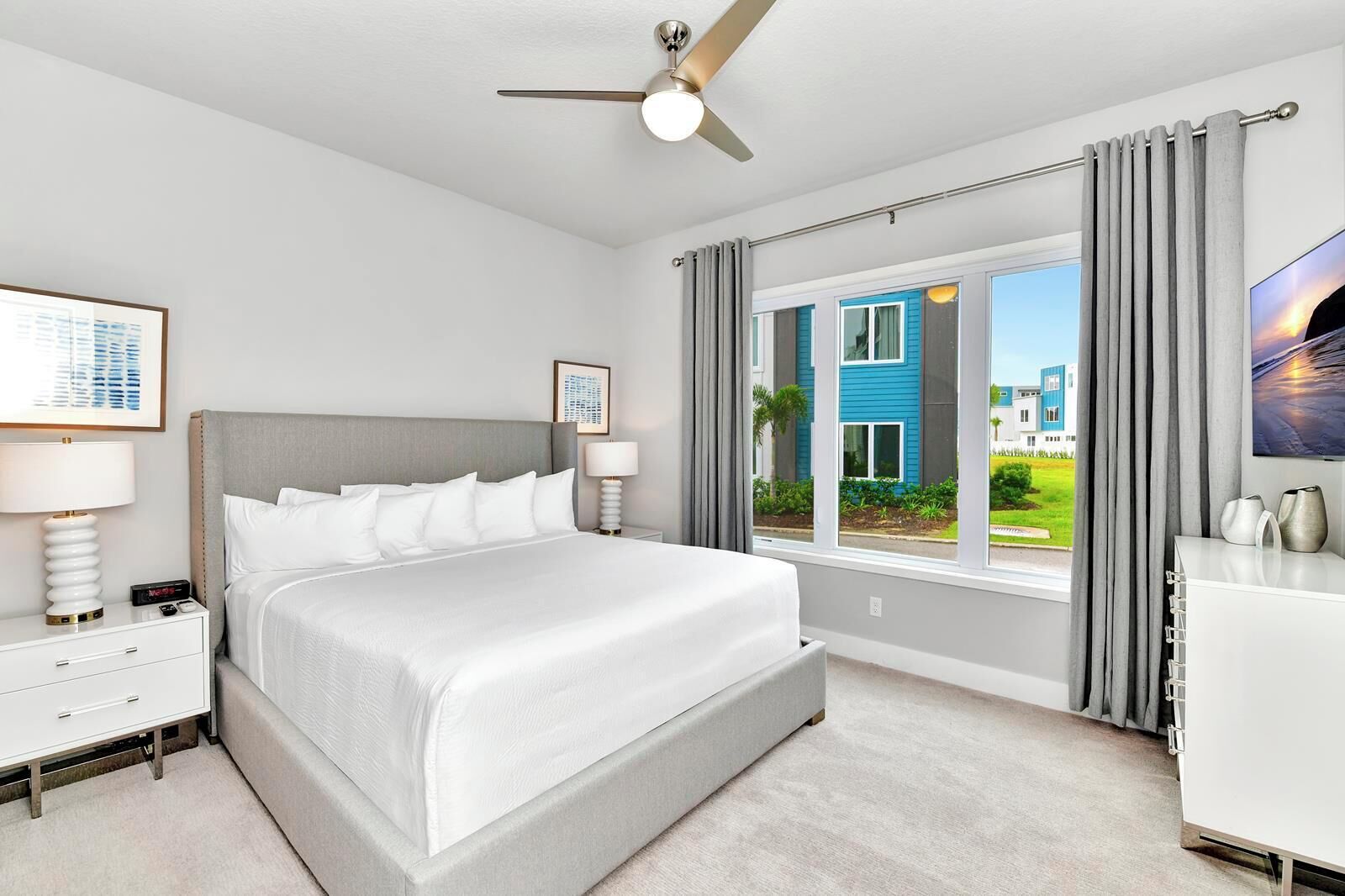 Una habitación de hotel con una cama grande, un ventilador de techo, una mesita de noche con lámpara, un aparador y una ventana con cortinas.