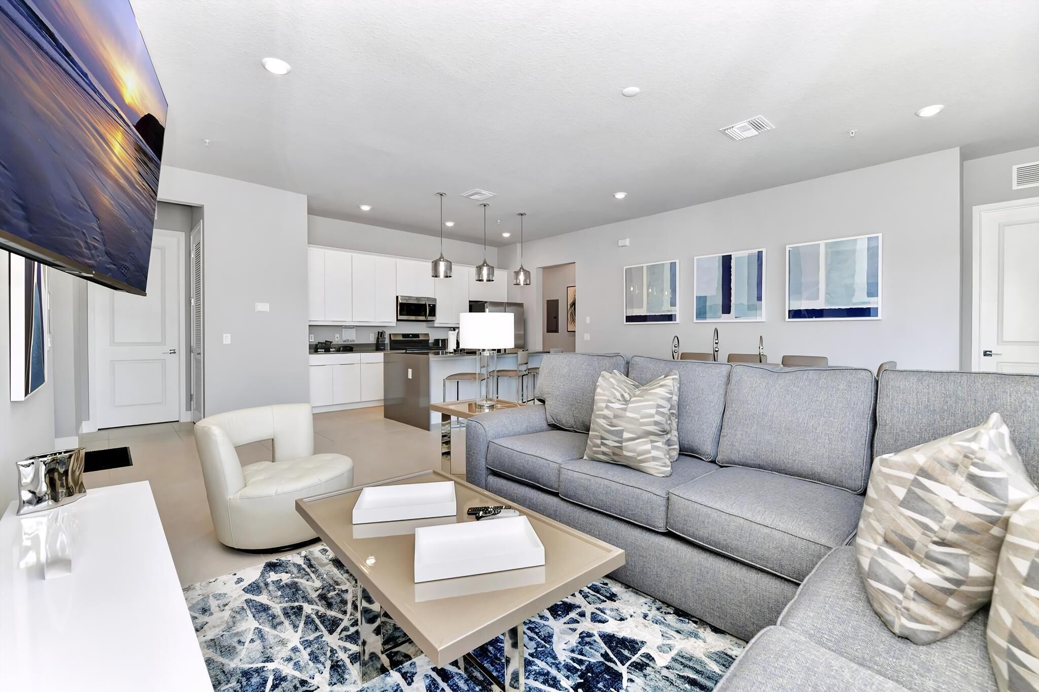 A modern living room with a grey sofa, white armchair, and a coffee table. There is a kitchen in the background with white cabinets and stainless steel appliances.
