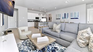 Ein modernes Wohnzimmer mit grauem Sofa, weißem Sessel und einem Couchtisch. Im Hintergrund ist eine Küche mit weißen Schränken und Edelstahlgeräten zu sehen.