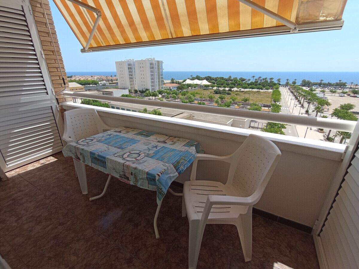 Ein Balkon mit Tisch und Stühlen, ein gestreifter Markise und Blick auf das Meer und Gebäude.