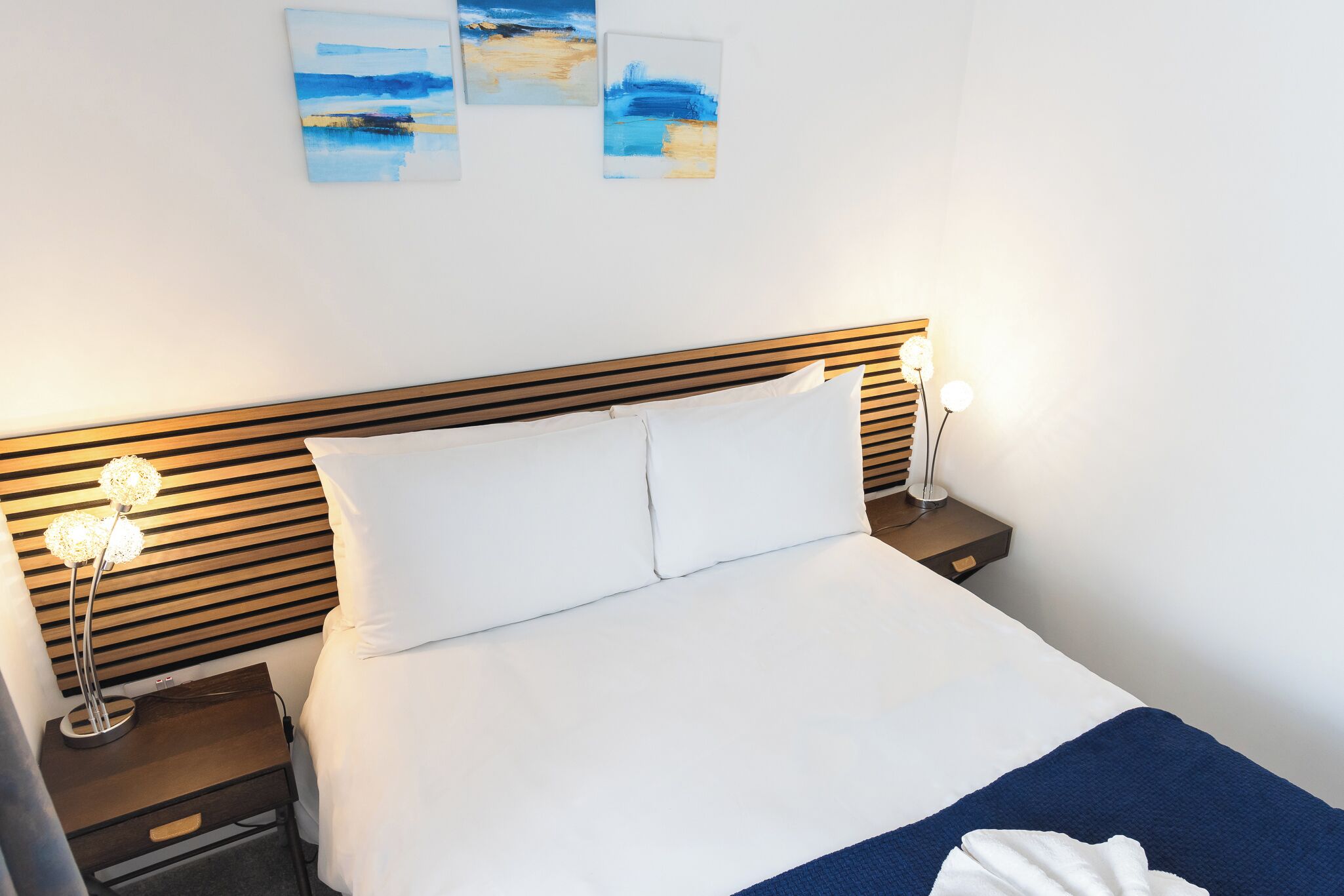 Ein ordentlich bezogenes Bett mit weißen Laken und einer blauen Decke, ein hölzernes Kopfende sowie Nachttischlampen auf einem Beistelltisch.