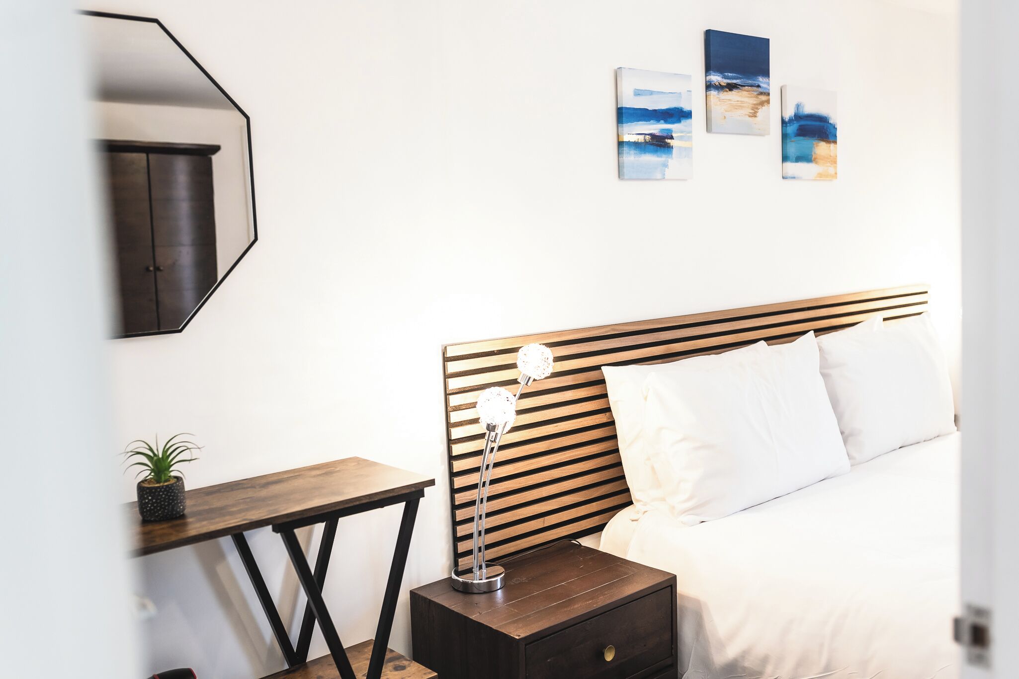 A neatly arranged bedroom with a wooden headboard, a bedside table with a lamp, a small potted plant, and a nightstand.