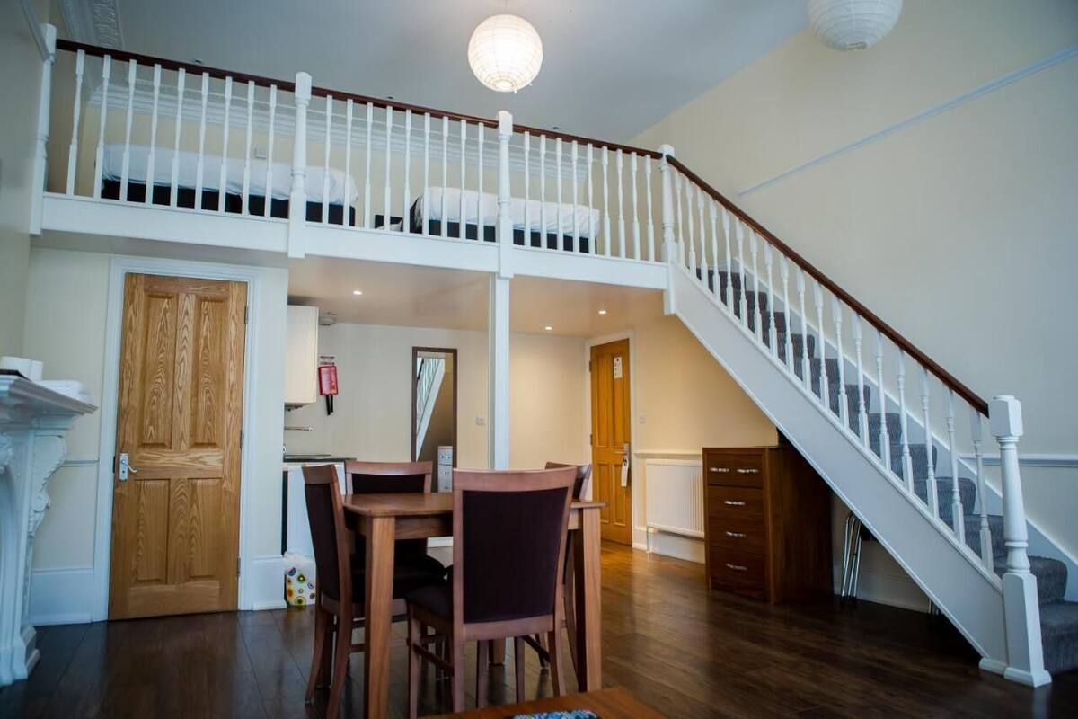 A spacious loft bedroom with a high ceiling, a wooden door, and a staircase leading to an upper level.