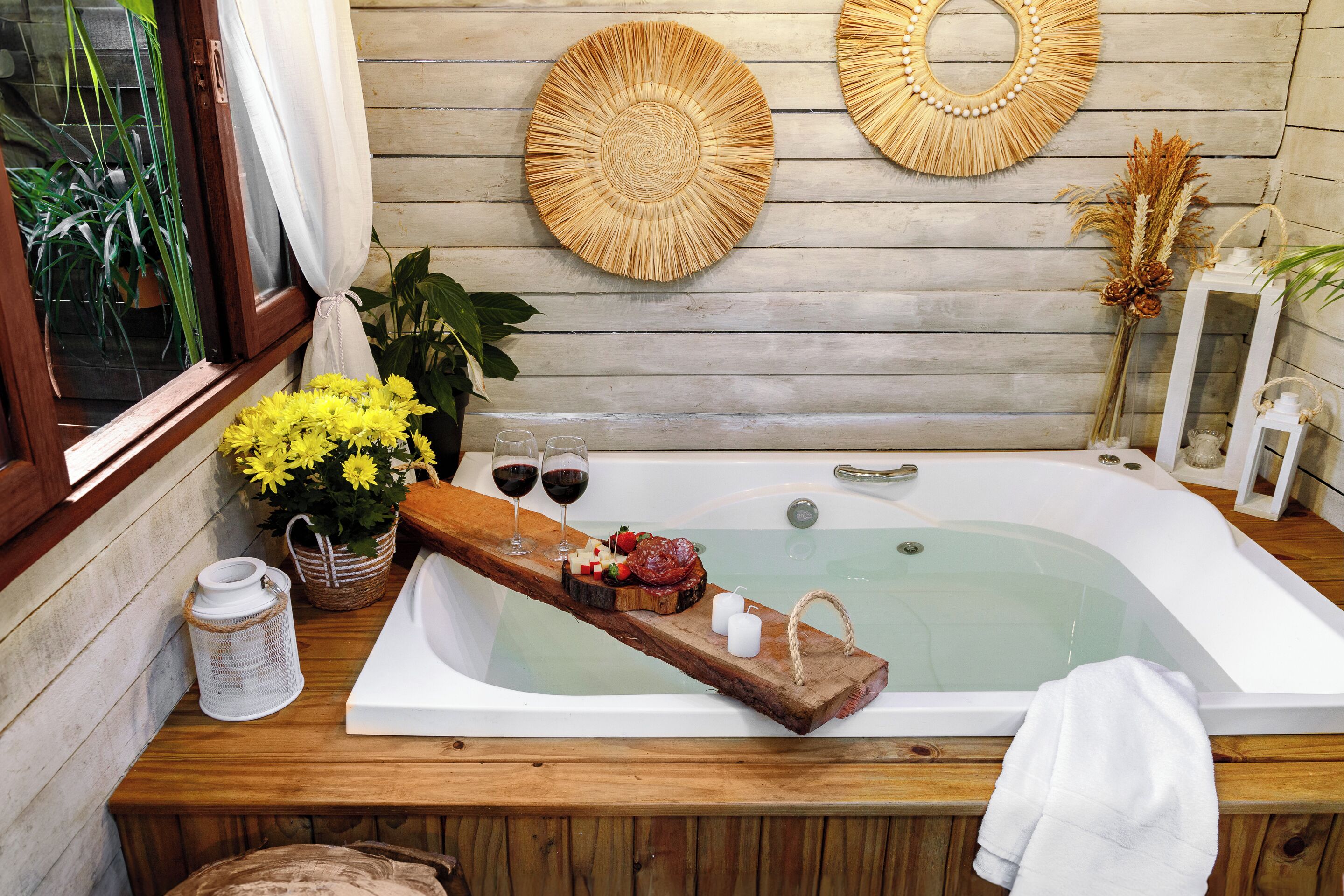 Une baignoire en bois avec un plateau en bois contenant des en-cas et deux verres de vin rouge, le tout entouré de plantes en pot et d’objets décoratifs.
