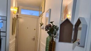 En smal hall med trägolv, en vit dörr längst in och en liten bänk. På en hylla finns dekorativa föremål, bland annat en vas med en växt och ett ljus.