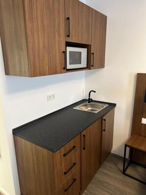 A compact kitchen with wooden cabinets, a black countertop, a stainless steel sink, and a microwave. - Munique Hotel Unterschleißheim (Unterschleissheim)