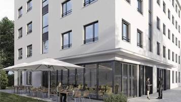 Ein modernes Gebäude mit flacher Fassade, zahlreichen Fenstern und einer überdachten Außenbereich mit Tischen und Stühlen.