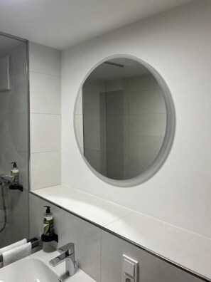 A modern bathroom with a round mirror, a white sink, and a shower area. - Munique Hotel Unterschleißheim (Unterschleissheim)