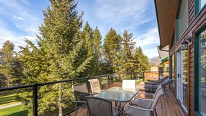 A wooden deck with a glass table and chairs, overlooking a forested area with tall trees and a clear sky.