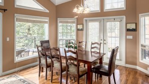 A dining area with a wooden table and chairs, large windows, and a view of the outdoors.