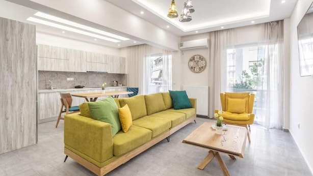 A modern living room with a sofa, a coffee table, and a yellow armchair. There is a kitchen area in the background with cabinets and a dining table. - Afroditi Suite (Nea Smirni)