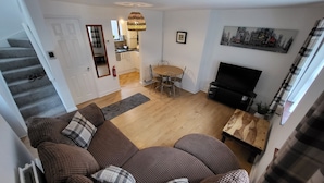 A living room with a brown sofa, a dining table with chairs, a TV, and a staircase leading to another room. - Heart of Lincoln City, 3 Bedrooms, Sleeps 4 Guests (Lincoln)