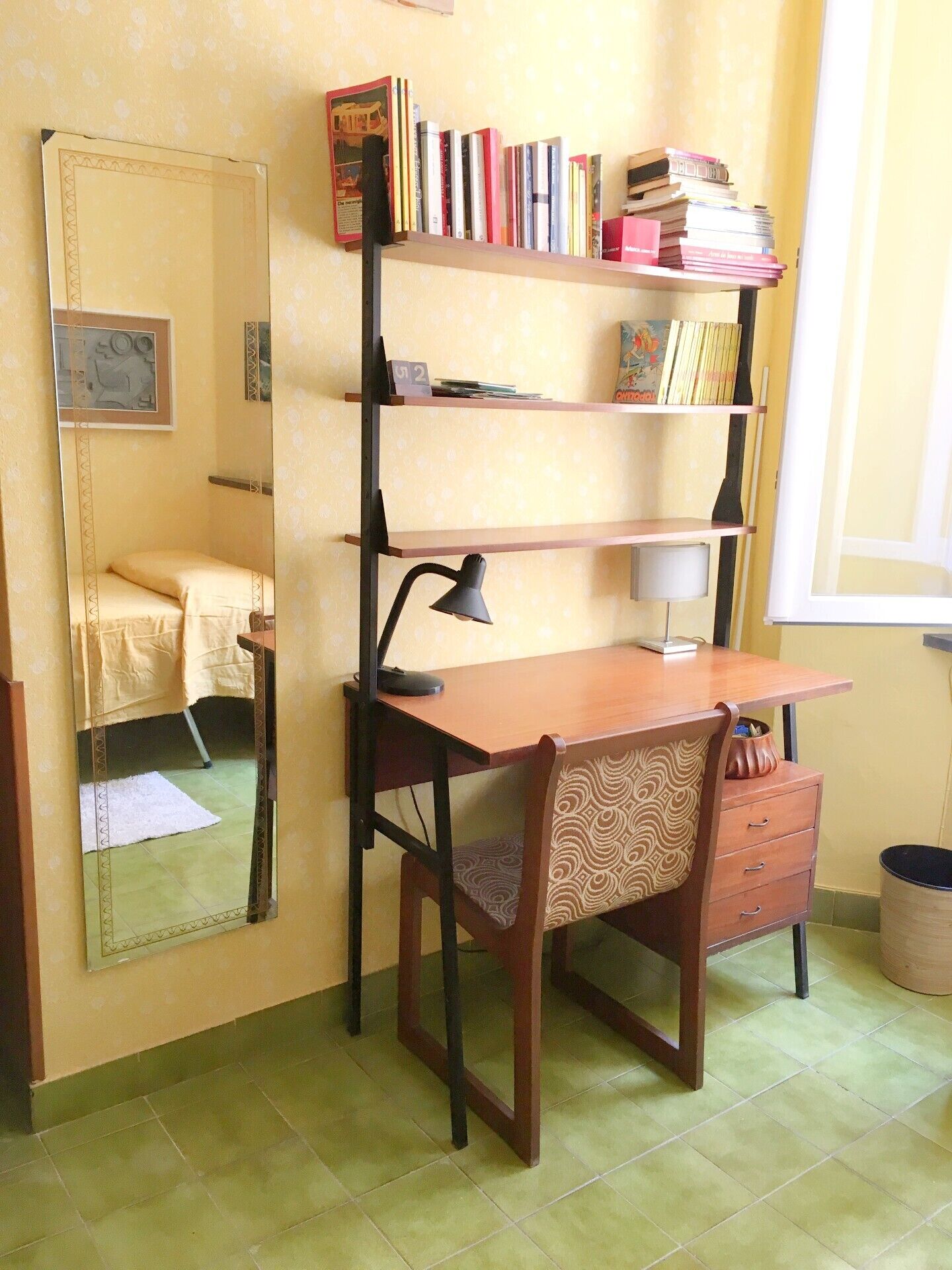 Una habitación compacta con un escritorio de madera, una silla, un espejo, una estantería y una ventana.