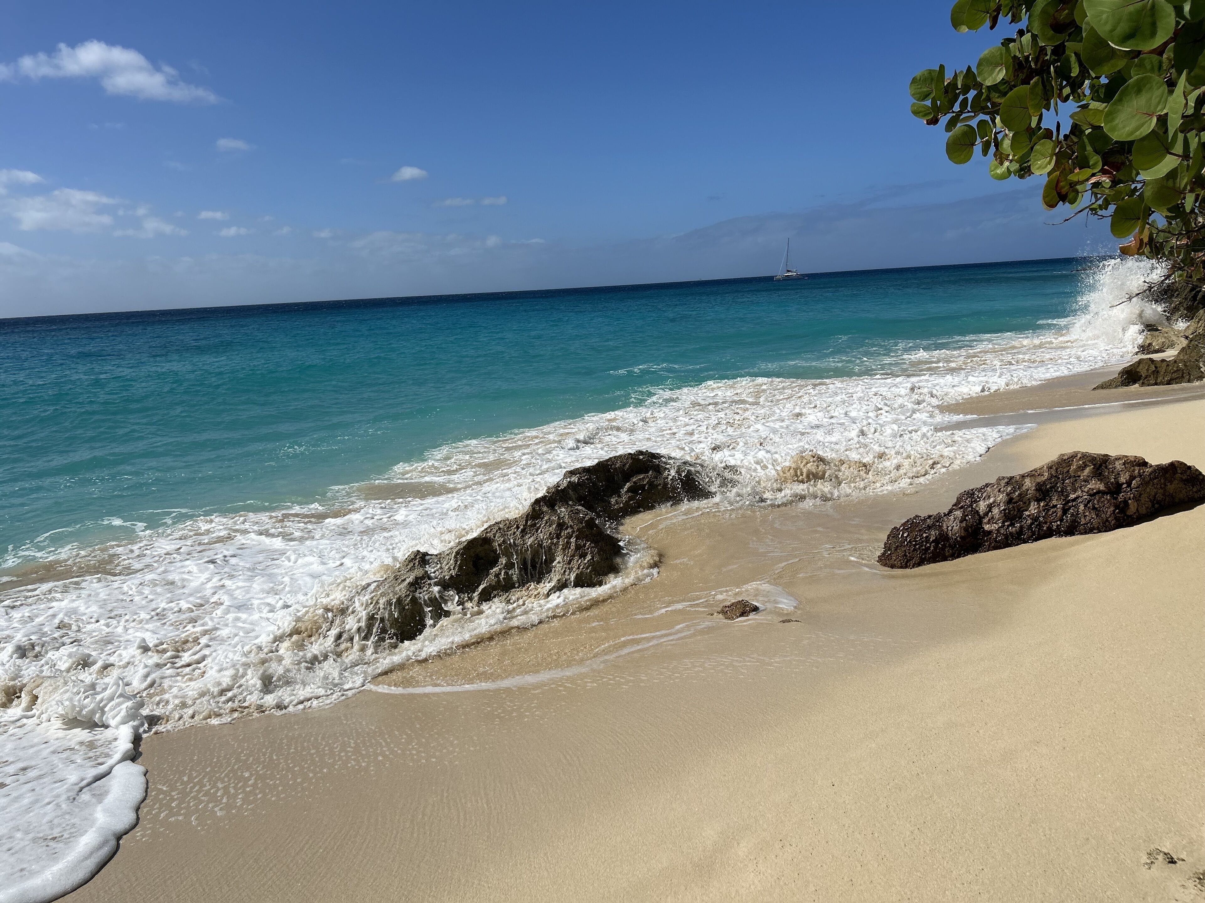 Une plage bordée d’une eau turquoise et limpide, un voilier à l’horizon et des affleurements rocheux.