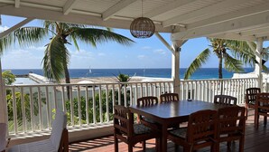 A covered outdoor seating area with a view of the ocean, featuring a wooden table and chairs, a white railing, and palm trees. - Holiday House in beautiful Cupecoy (Cupecoy Beach Club)