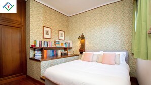 Ein Schlafzimmer mit einem Bett, einem Nachttisch mit Büchern, einer Nachttischlampe und blumigem Wandpapier.