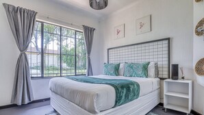 A bedroom with a large bed, a bedside table, a window with curtains, and a wall-mounted shelf.