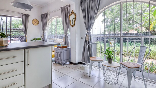 A bright kitchen with white cabinets, a grey countertop, and a hanging pendant light. There is a window with grey curtains and a view of greenery outside.