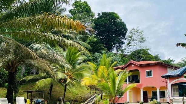 A pink house with a covered porch, surrounded by lush greenery and palm trees. - Studio Avec Vue sur Jardin (Mpolongwé)
