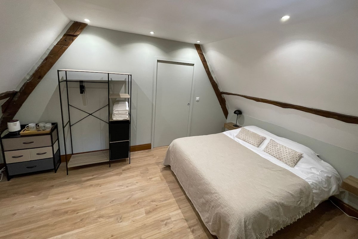 Un dormitorio con techo inclinado, una cama con colcha blanca, una mesita de noche con lámpara, un armario y suelo de madera.