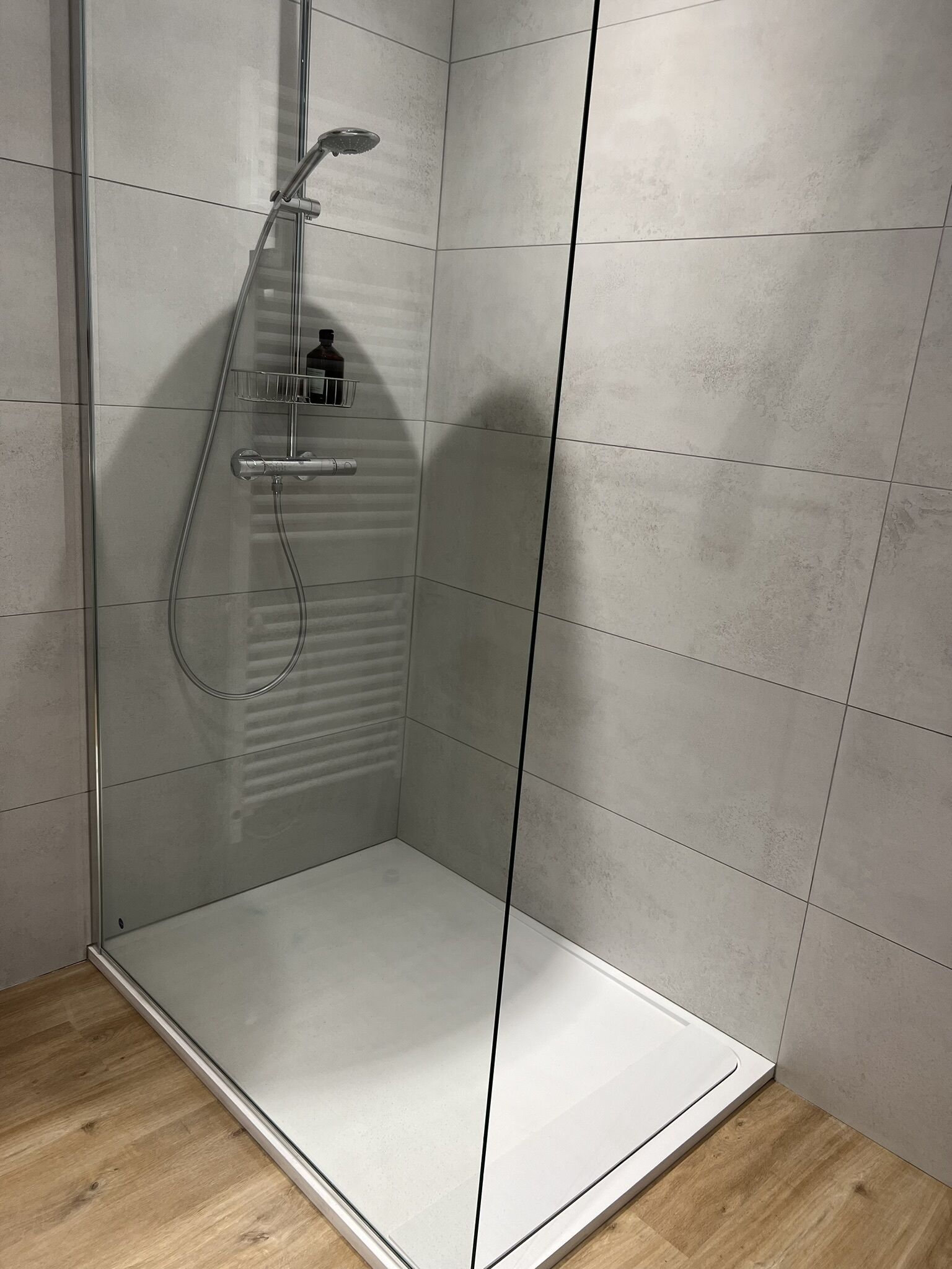 A glass-enclosed shower with a handheld showerhead, a bottle of shampoo, and a towel rack.