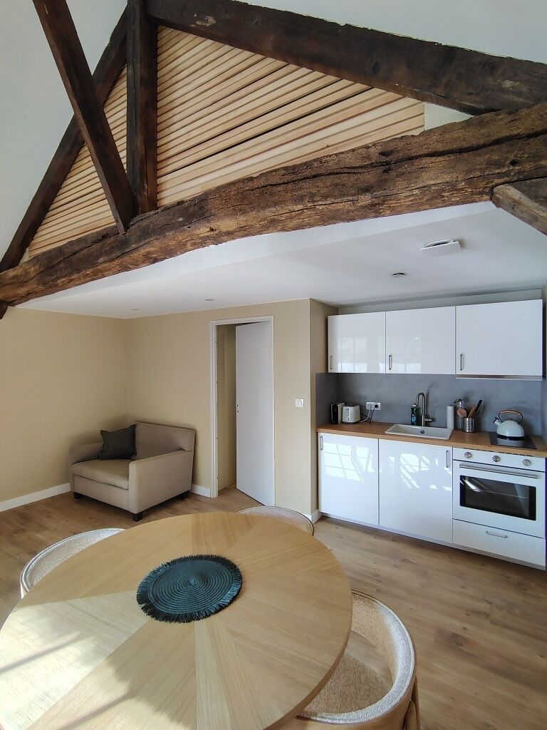 A room with a wooden dining table, a sofa, a kitchen with white cabinets, and exposed wooden beams on the ceiling.