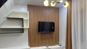 A modern living room with a flat-screen TV mounted on a wooden panel wall, a white entertainment unit with shelves, a brown sofa, and a ceiling light fixture with multiple bulbs. - PearlDaisy Butuan Transient Home Philippines (Butuan City)