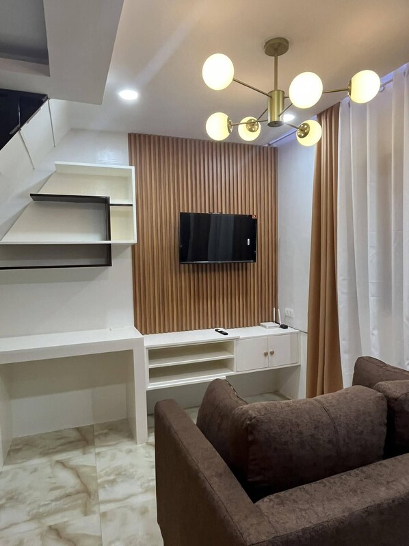 A modern living room with a flat-screen TV mounted on a wooden panel wall, a white entertainment unit with shelves, a brown sofa, and a ceiling light fixture with multiple bulbs. - PearlDaisy Butuan Transient Home Philippines (Butuan City)