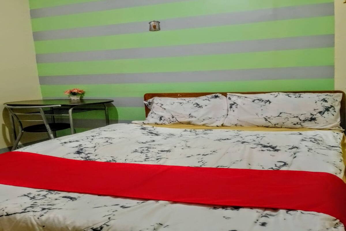 A room with a bed, a desk, and a chair. The bed has a red blanket and white sheets with a black pattern. The desk has a small table with a flower vase. The walls have green and gray stripes.