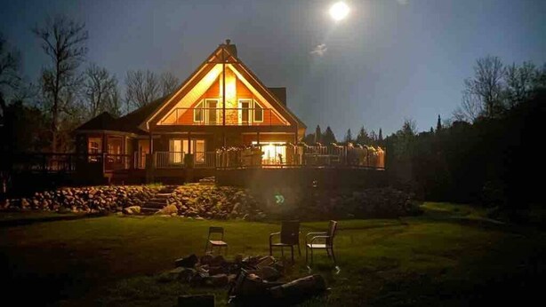 A large, two-story house with a prominent front porch and a well-lit exterior. - 5 Bedroom Waterfront Lake House  (Bonfield)