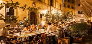 An outdoor dining area with tables and chairs, surrounded by buildings with arched windows and a staircase.