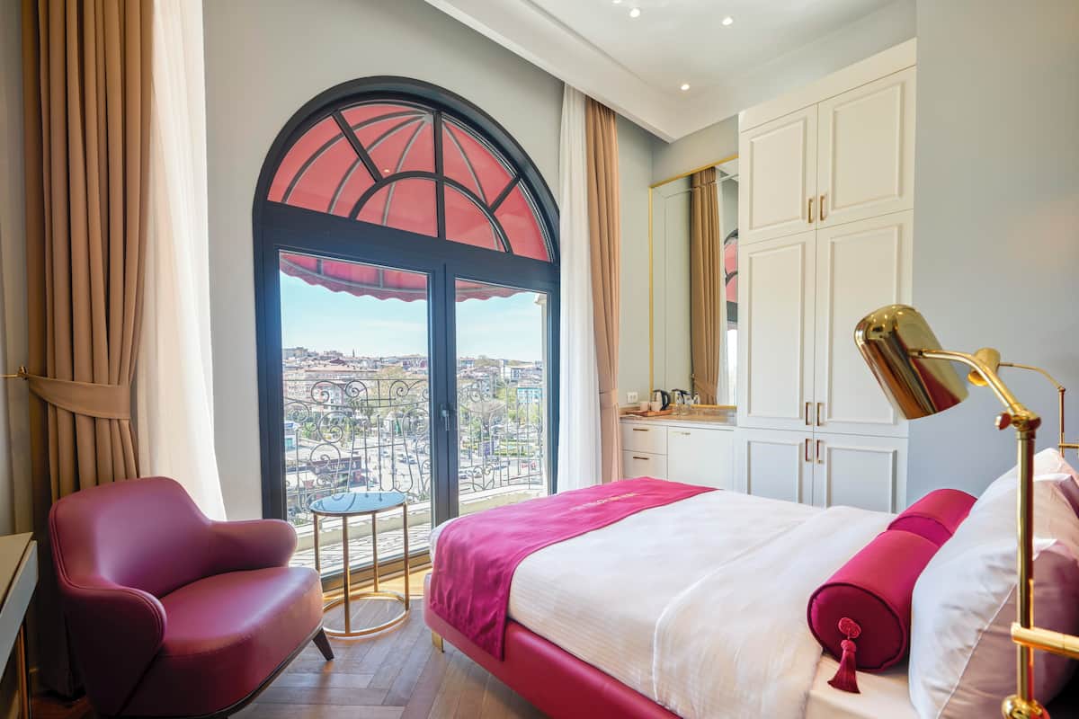 A hotel room with a large bed, a desk, a chair, a small table, and a balcony with a view.