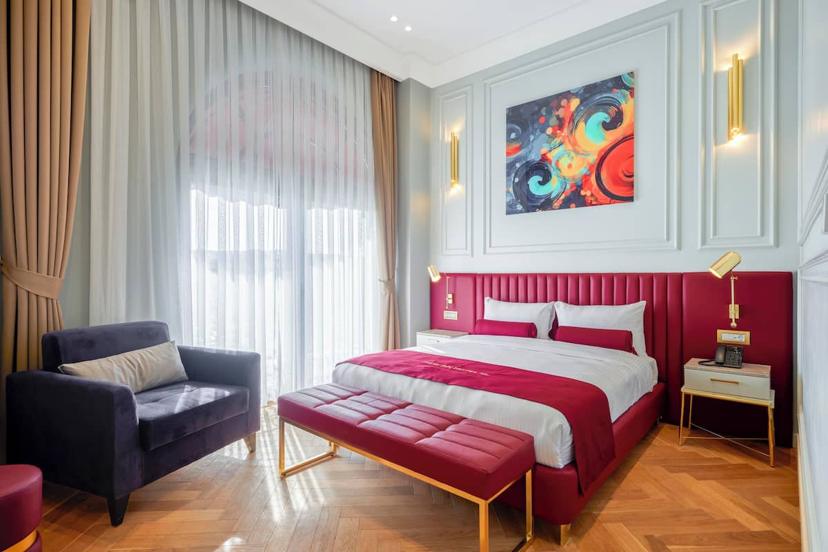 A modern hotel room with a large bed, a red upholstered bench, a dark armchair, and a nightstand with a lamp.