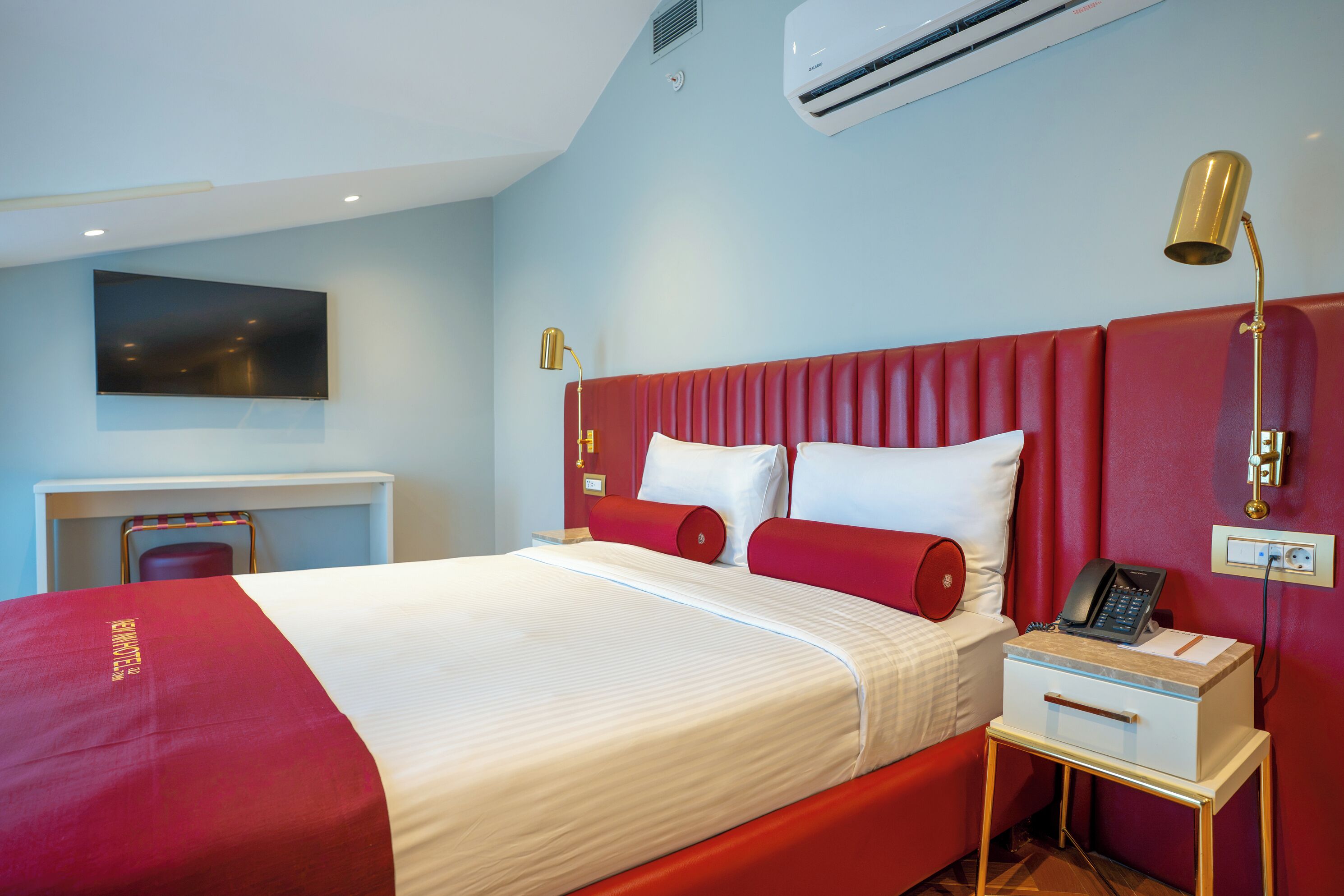 A hotel room with a bed, a bedside table with a lamp, a TV, and an air conditioning unit.