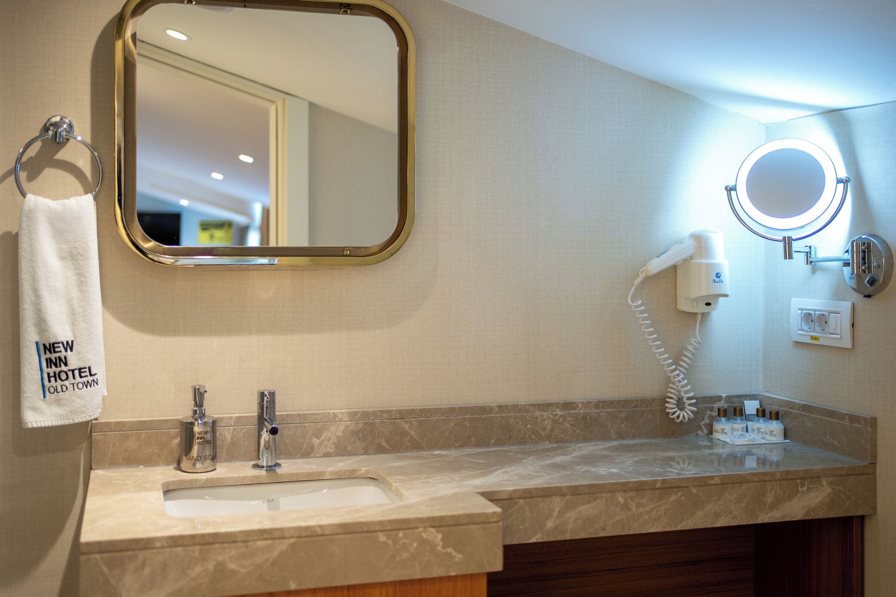 A bathroom with a sink, a mirror, a hand soap dispenser, a hairdryer, and a towel hanging on a hook.