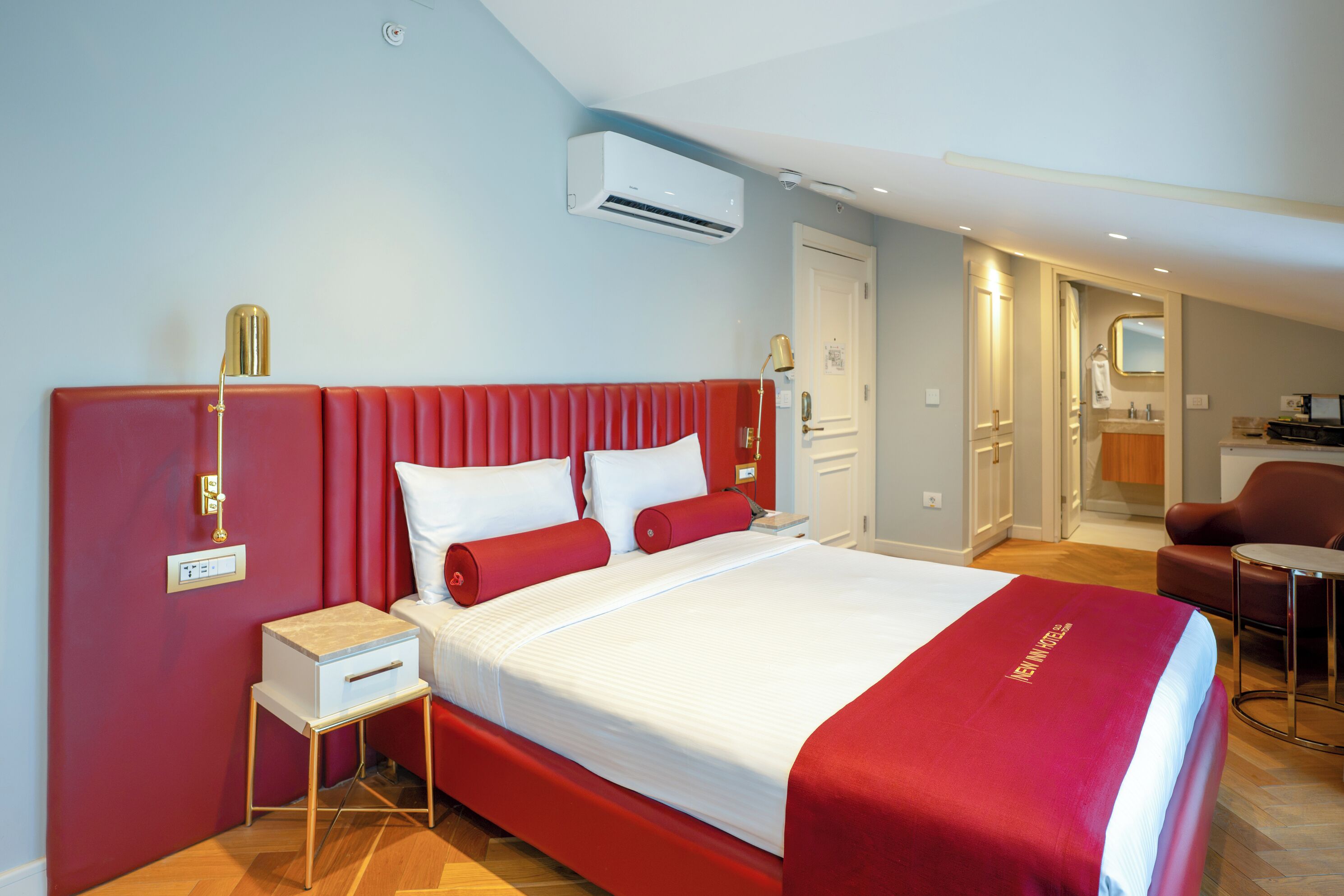 A hotel room with a large bed, a bedside table, a nightstand, a red upholstered headboard, an air conditioning unit, a door, a mirror, a chair, and a small table.