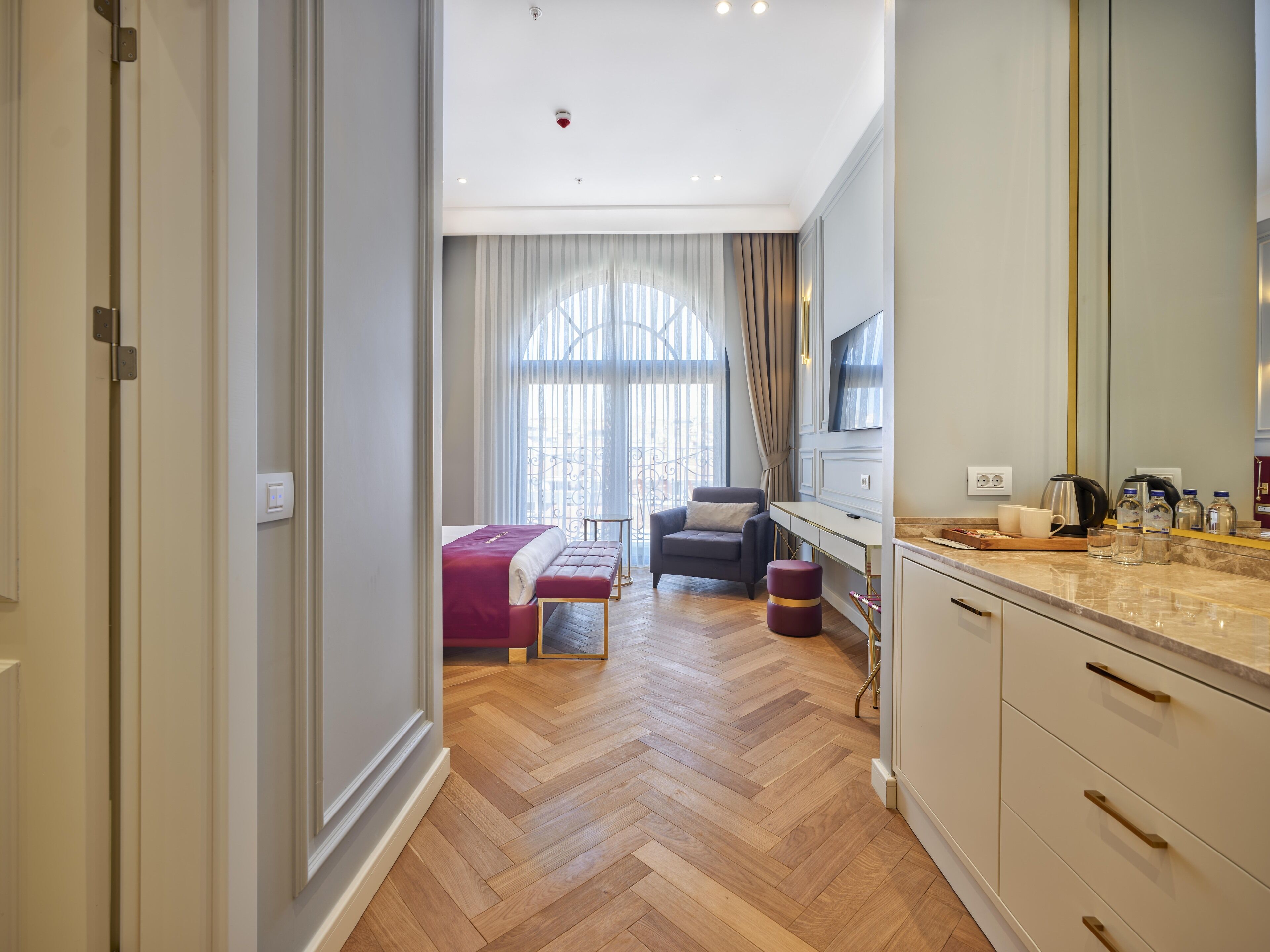 Una habitación de hotel moderna con piso de madera en patrón espiga, una cama con cabecera roja, un área de descanso con un sillón y reposapiés, una cocina con encimera de mármol y un ventanal con cortinas.