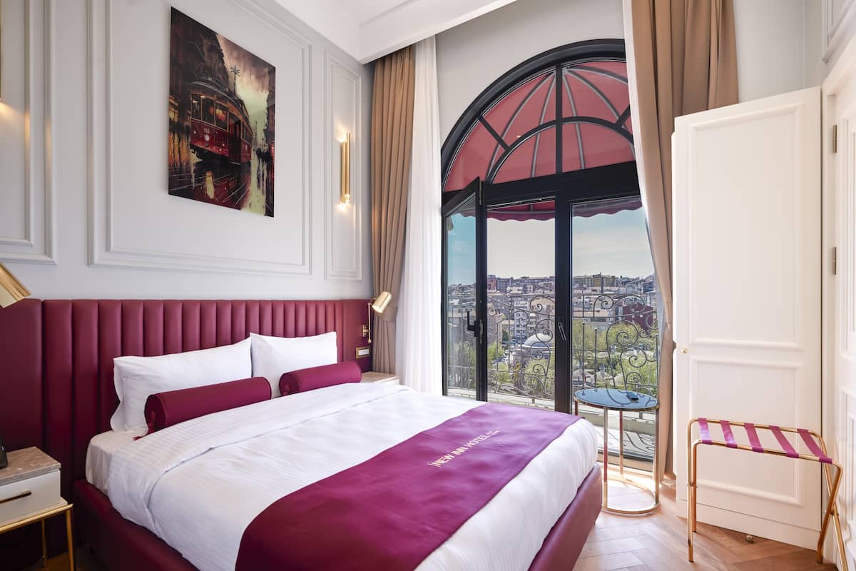 A hotel room with a large bed, a city view through a window, a painting on the wall, and a balcony with a table and chairs.