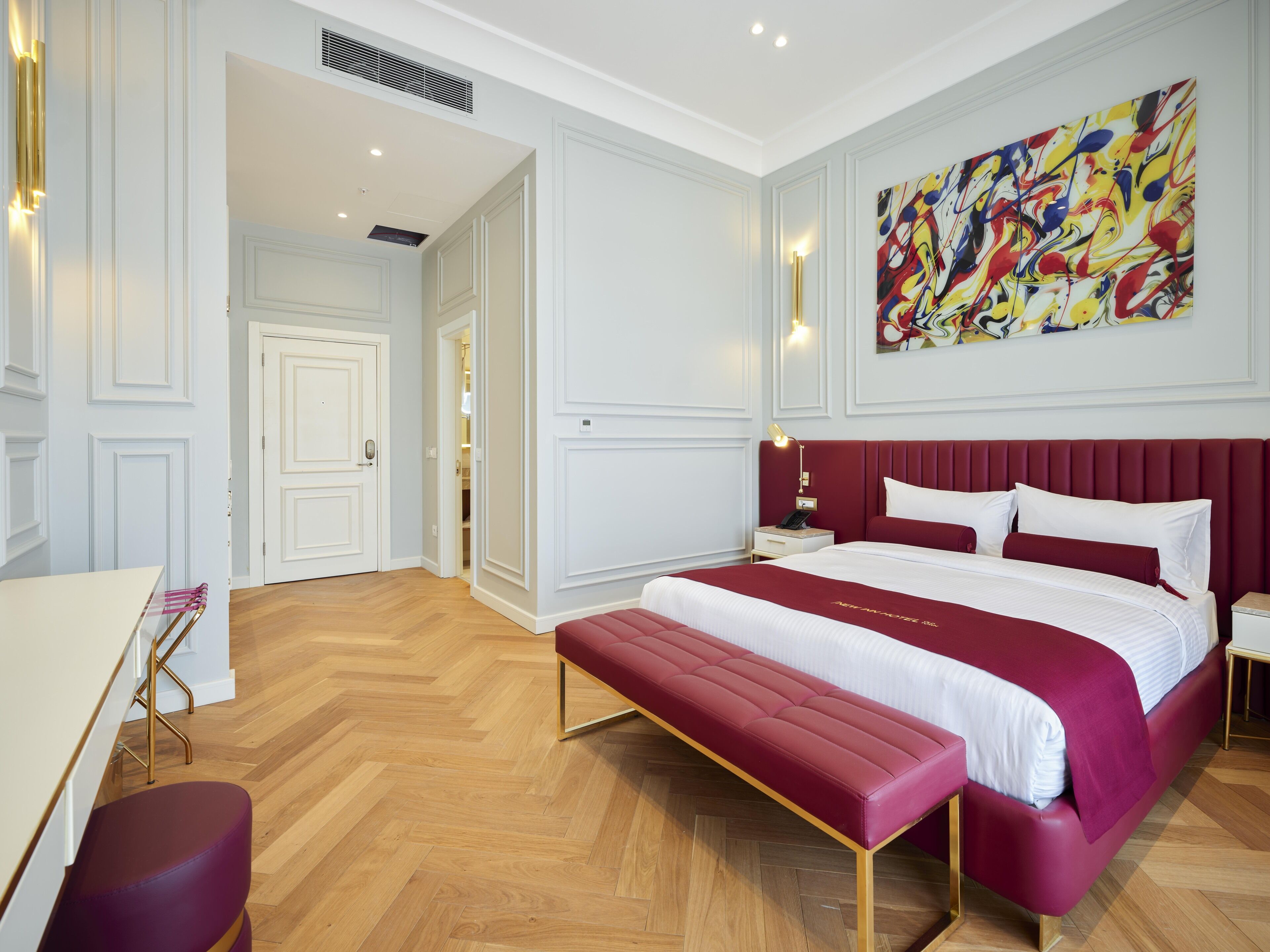Une chambre d’hôtel moderne avec un grand lit, une banquette, un bureau et une peinture abstraite aux couleurs vives accrochée au mur.