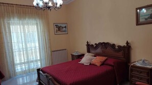 Un dormitorio con una cama grande, una araña, un radiador, un aparador de madera y una ventana con cortinas.